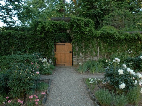 A garden gate surrounded by stone wall and flowering plants in foreground.