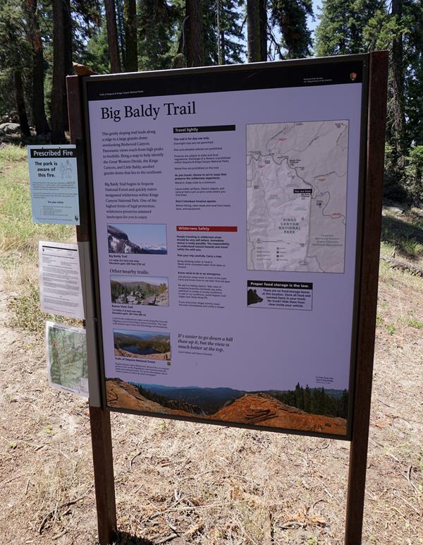 A metal framed sign sits i front a dirt path leading to a stand of trees