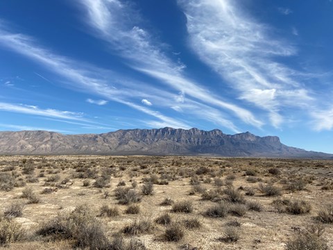 A desrt mountain range looms over a low desert foreground with sand dunes in the distance