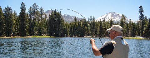 A man holds a fly fishing on a blue lake backed by conifer trees and volcanic peaks.
