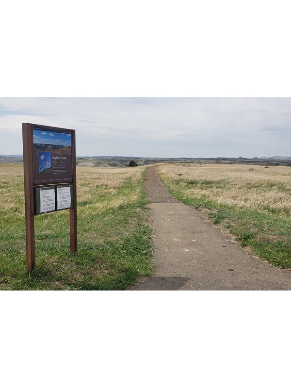 A trailhead sign next to a paved trail in a grassy area. Buttes are visible on the horizon.