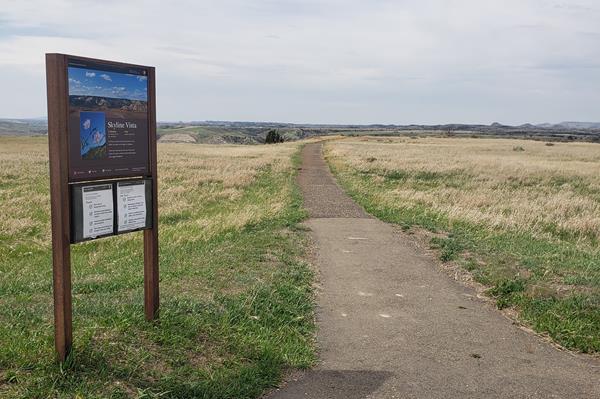 A trailhead sign next to a paved trail in a grassy area. Buttes are visible on the horizon.