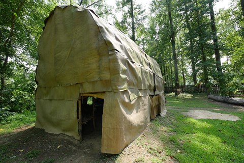 A Native American longhouse sitting in a grassy area under trees.