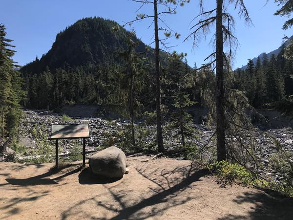 A wayside panel in a metal frame overlooks a wide, rocky riverbed and forested hills..