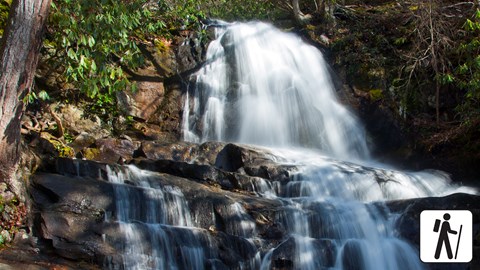 A two-tiered waterfall framed by greenery. Hiker icon in lower right corner.