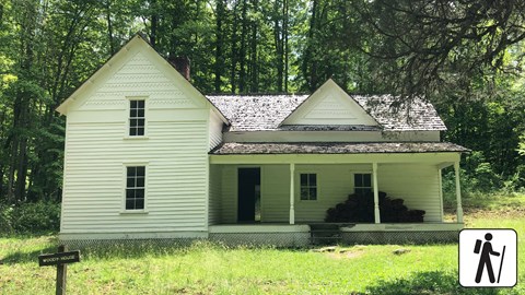 A white historic home in a grassy field with forest behind it. A wooden sign labels the home.