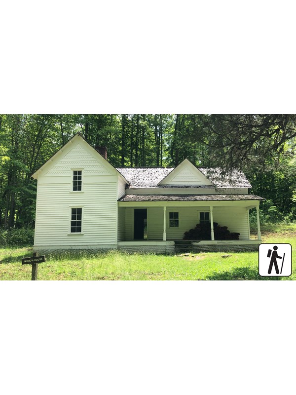 A white historic home in a grassy field with forest behind it. A wooden sign labels the home.