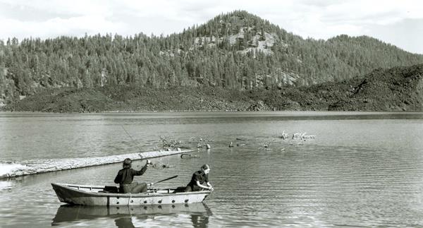 Two women fishing from a row boat on a lake backed by a lava flow and a volcanic peak.