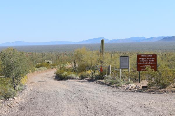 Dirt road leading into the desert. At the entrance is a gate and sign about border activity.