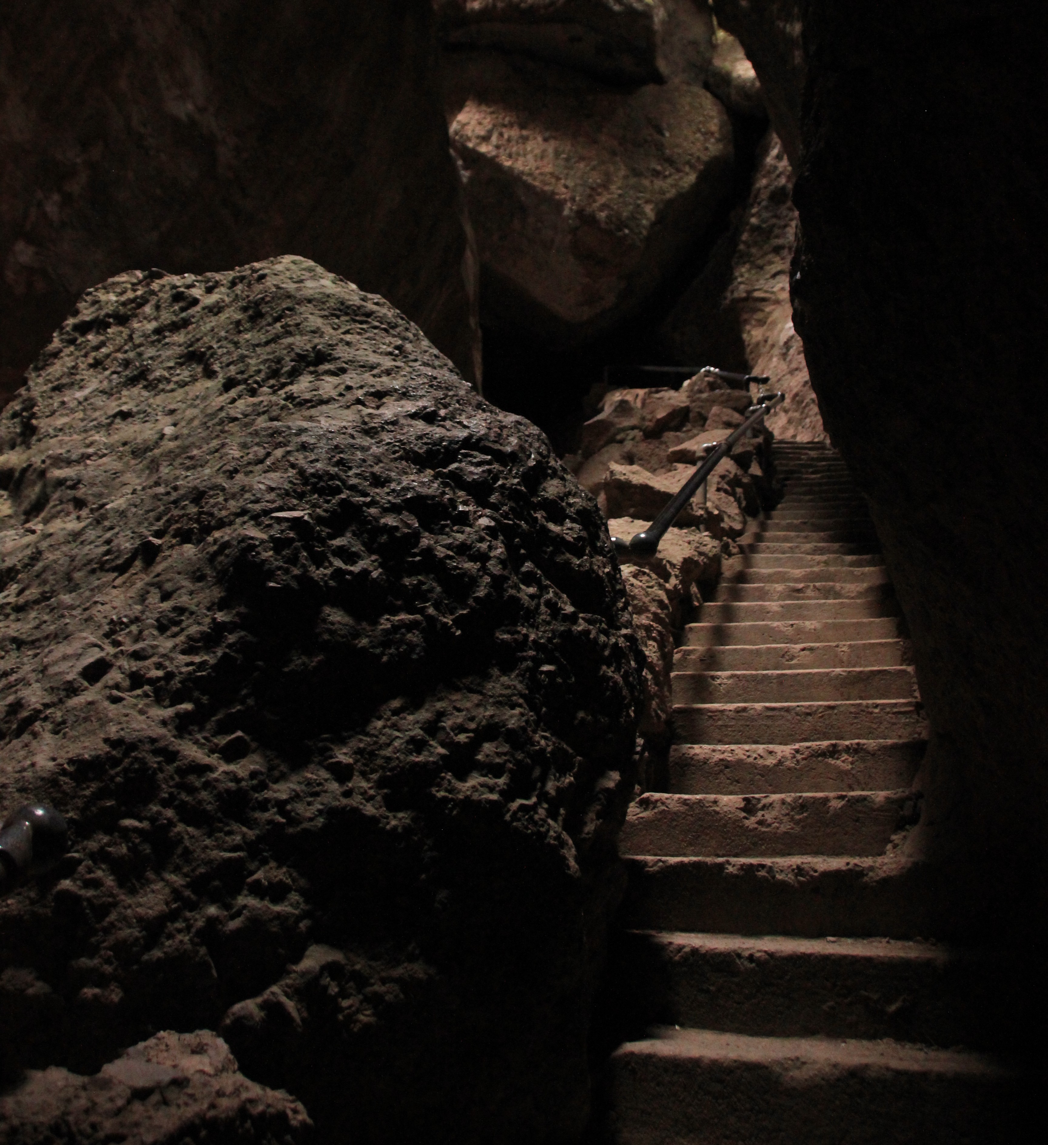 View of a large boulder and rock carved steps adjacent a handrail underneath a rock roofed canyon.