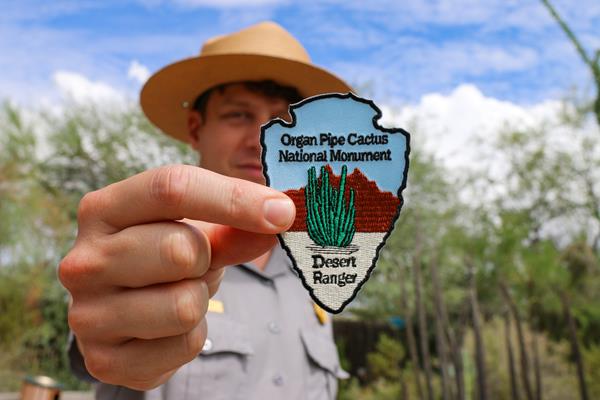 Ranger holding a Desert Ranger patch up to the camera. Native cacti are in the background.