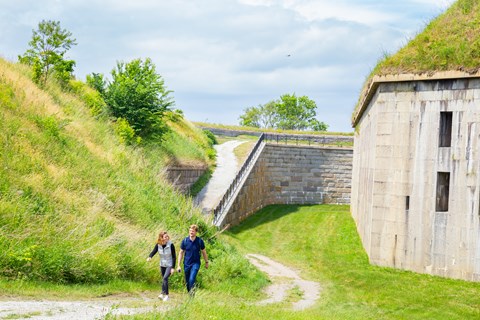 Two people walking on a trail with a fort's granite wall to the left of them.