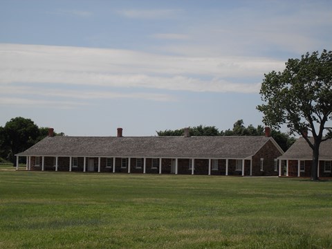 Long distance view of one-story sandstone building with covered porch held up by white columns.