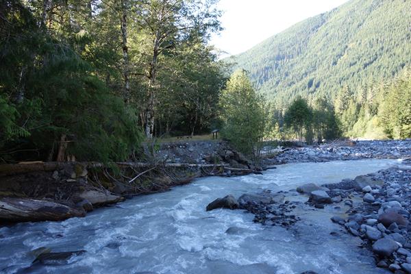 A wayside in a metal frame stands at the edge of a rocky river bed in a forested valley.