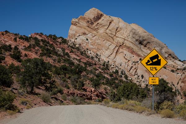 Dirt road that curves around a large rock. Road sign indicating steep incline ahead.