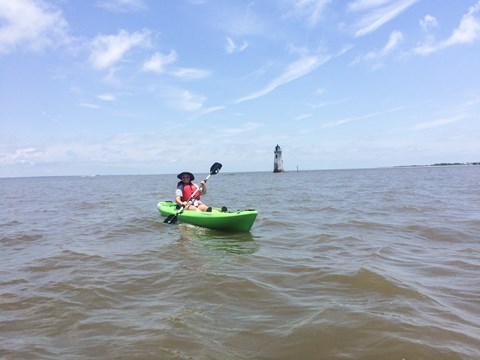 A woman in a green kayak with a lighthouse distantly behind.