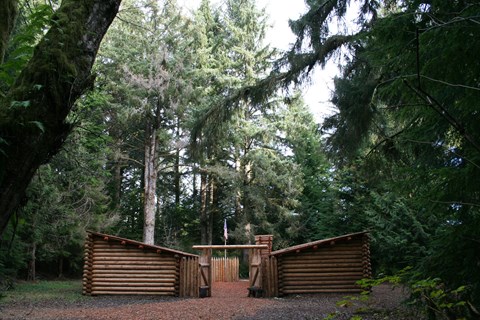 A log fort in a dense evergreen forest. Two parallel log structures with a flag in between.