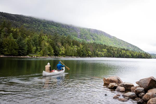 Two people, wearing jackets and hats, paddle a silver canoe in water surrounded by trees.