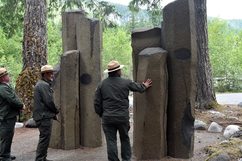 Three park rangers stand next to a stone memorial made of seven basalt columns