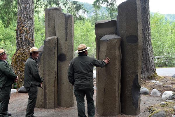 Three park rangers stand next to a stone memorial made of seven basalt columns