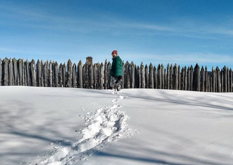 A person makes snowshoe tracks in front of a stockade.