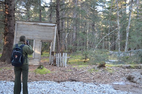 Woman in National Park Service uniform looking at remains of historic wooden building.