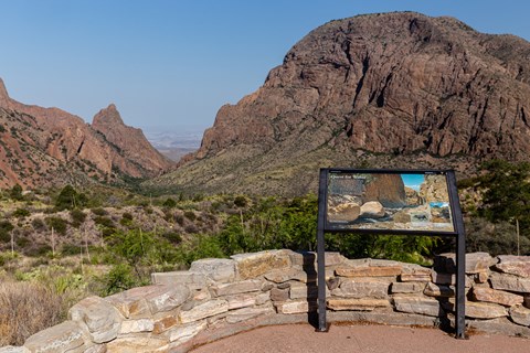 A paved trail and metal exhibit sign overlook a low basin surrounded by mountains.