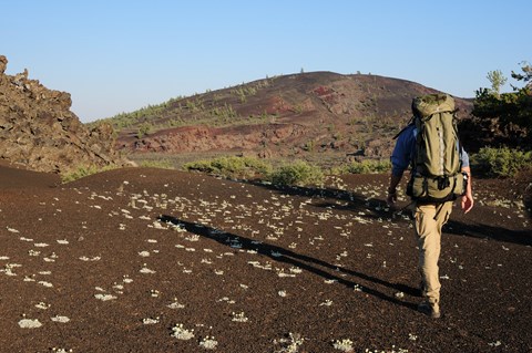 A hiker with a large backpack walks on a cinder trail toward a large volcanic cone.