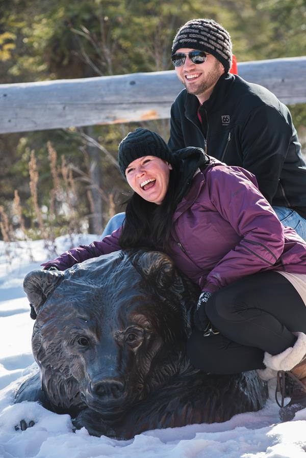 a man and woman smile for a photo while sitting atop a sculpture of a grizzly bear