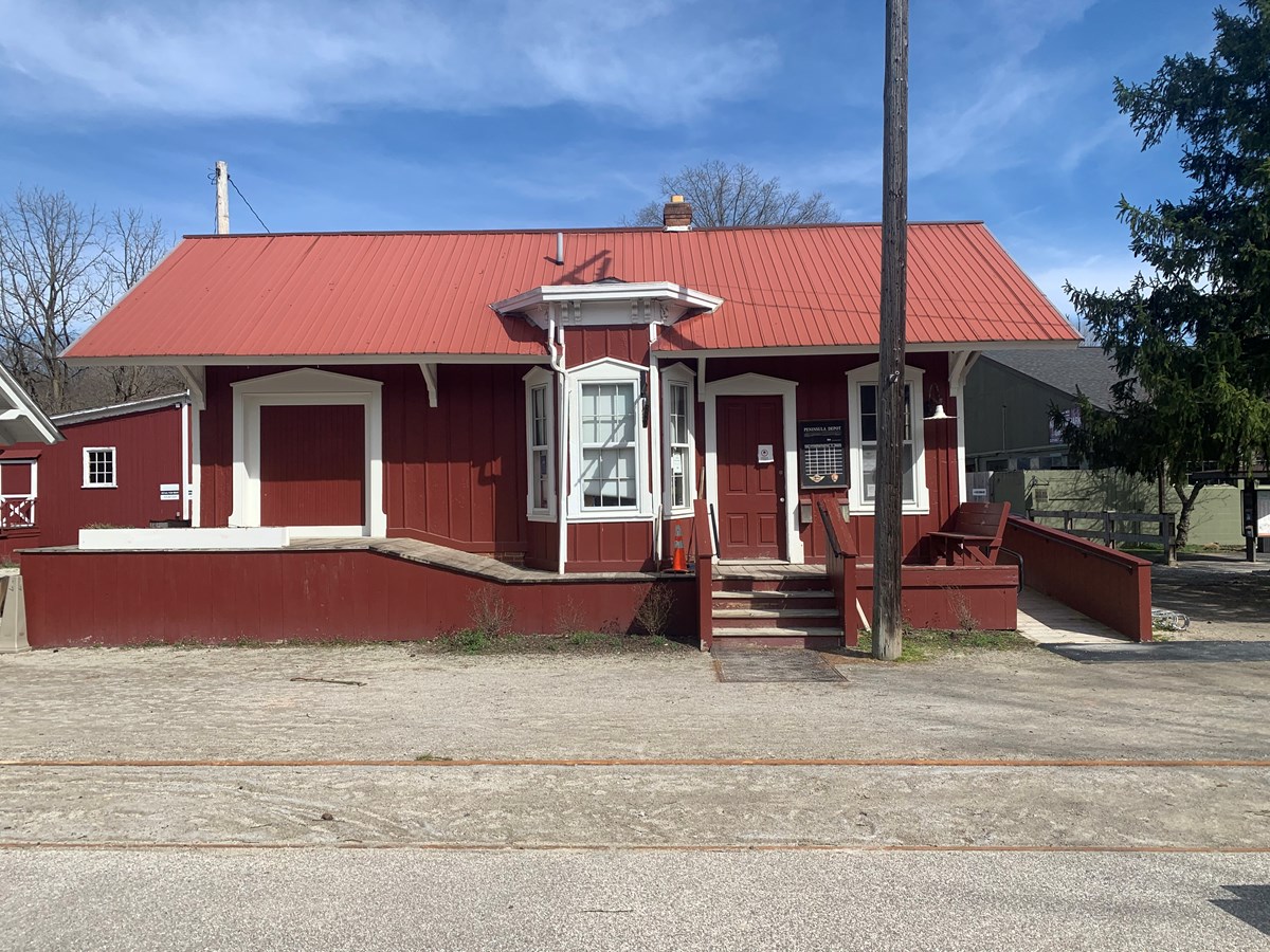 A red building with white trim; the depot has a platform, ramp, posted train schedule and bay window.