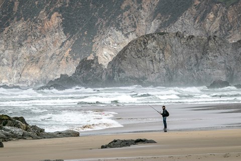 A man fishing on a sandy beach with waves washing in from the left & steep bluffs in the background.