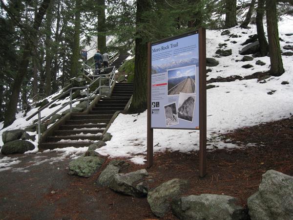 A trailhead sign is situated in the foreground. Stone stairs that summit Moro Rock in background.
