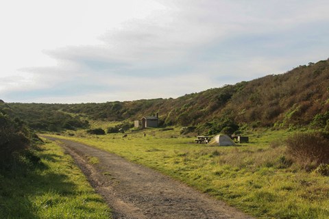 A small camping tent adjacent to a hillside on green grass, next to an outhouse and gravel path.