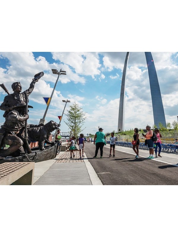 a group of people standing along a trail next to the Captain's Return statue in front of the Arch