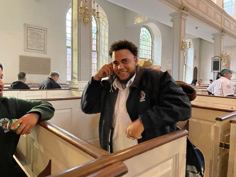a man holding up an audio tour set to his ear while standing in a pew.