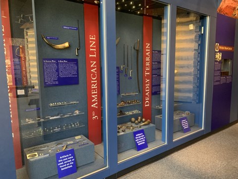 A museum exhibit with blue and red planning featuring weapons like musket balls & bayonets