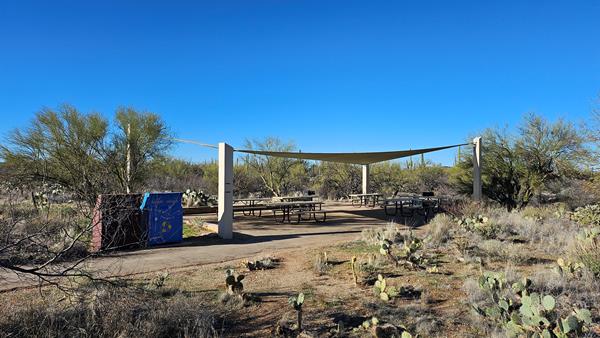 A large triangular shade structure covers six metal picnic tables. Two trash cans left of tables