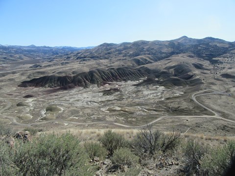 View of red and green striped hills from trail above, with green sagebrush and windy dirt road below