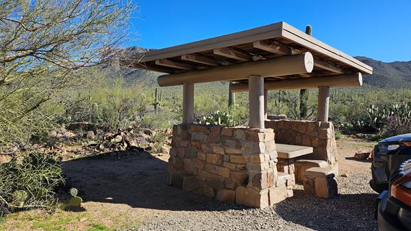 A rock and wood shade structure covers a stone picnic table, surrounded by a lush desert landscape.