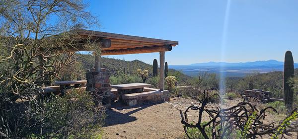 A large wood and stone shade structure covers two stone tables (left). To the right spans a valley.