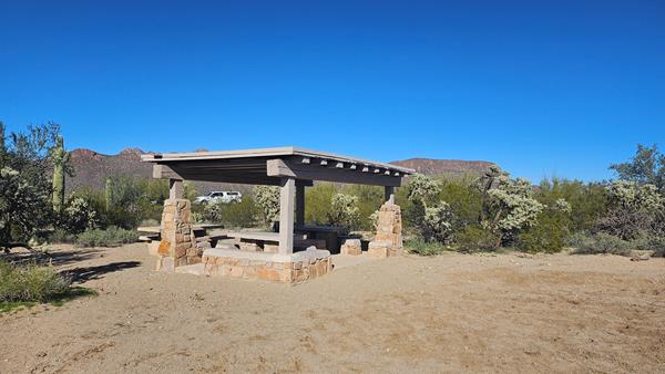 A large stone and wood structure shades two stone tables, with another stone table is uncovered.