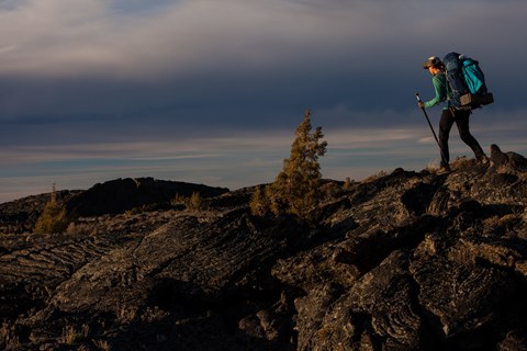 A hiker with a large backpack traverses a lava field.