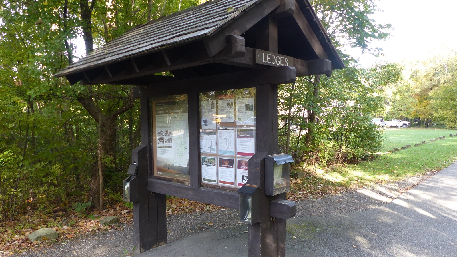 A two sided bulletin board along a paved road; a “Ledges” sign just below the peaked roof.