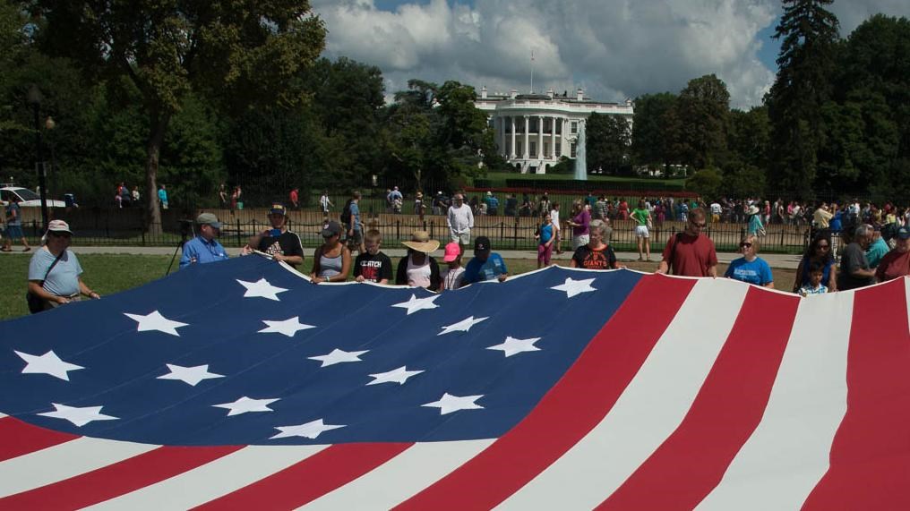 A crowd of people holding a large Star-Spangled Banner replica flag in front of the White House