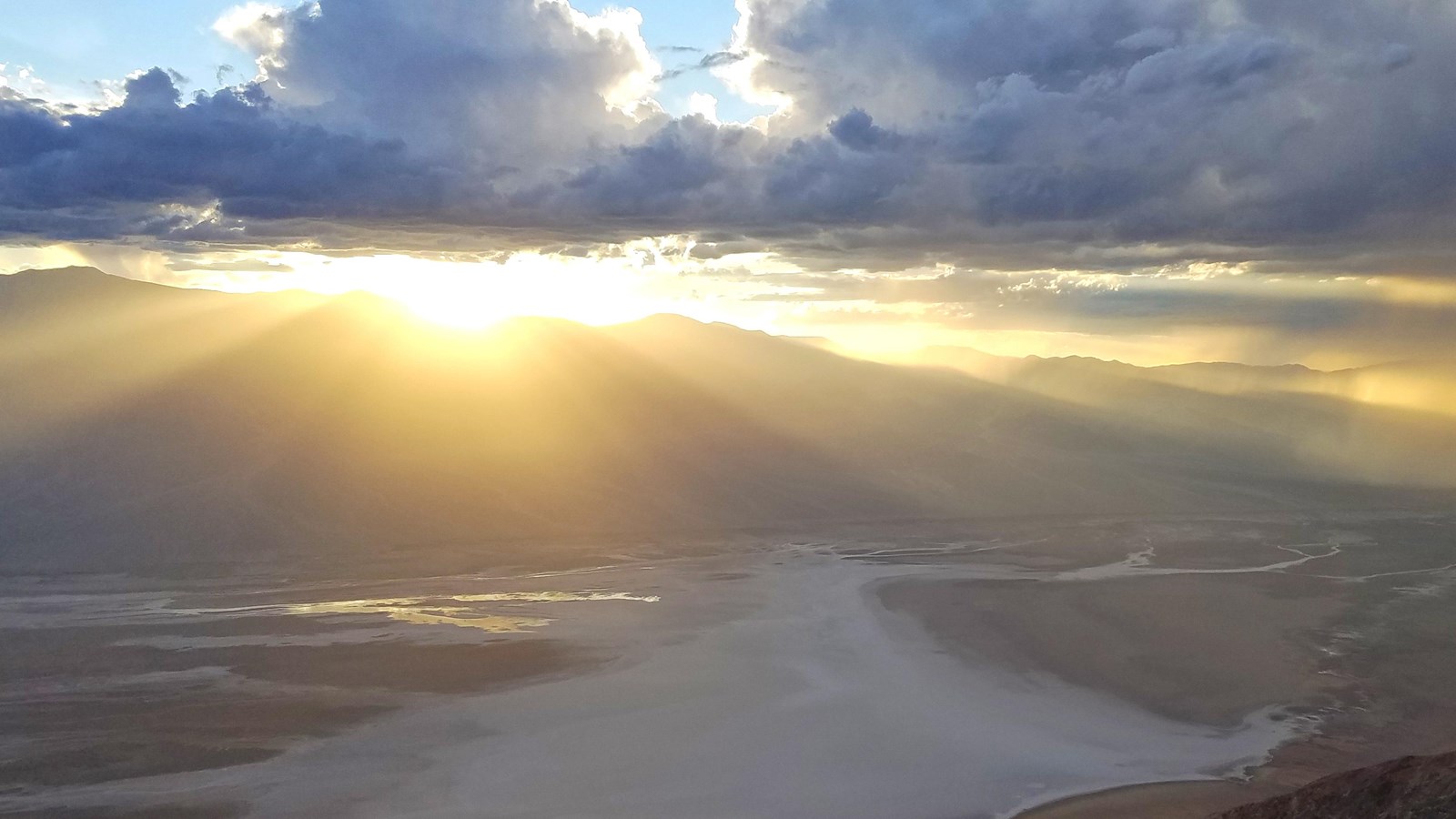 Shafts of light stream through clouds over a dark mountain range and swirling white salt flats.