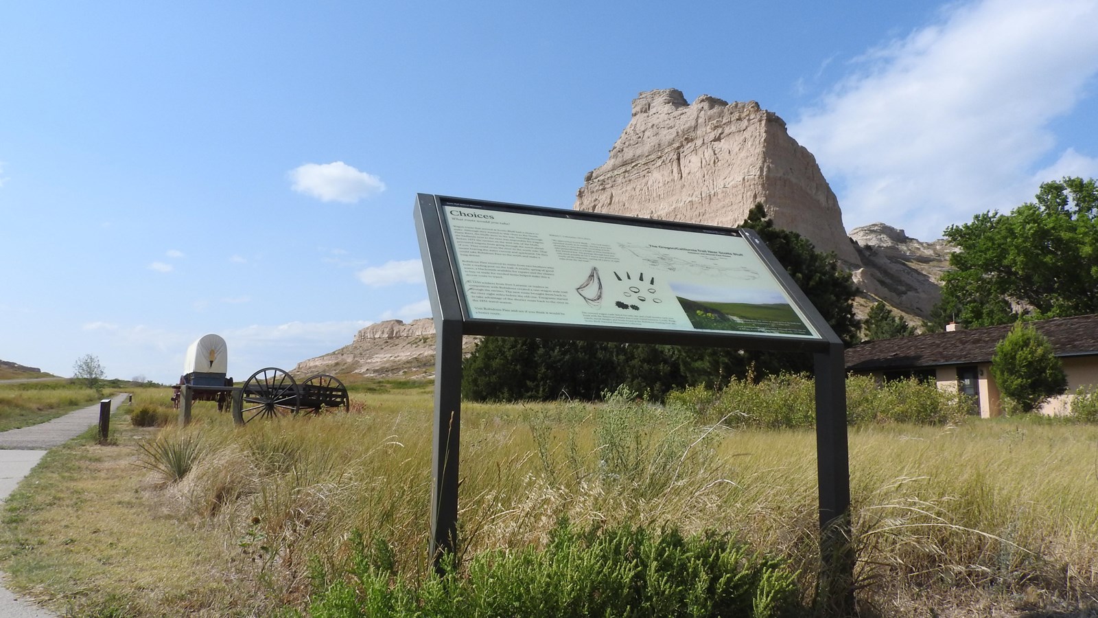 A wayside sign sits to the right hand side of a paved trail with a view of a sandstone bluff. 