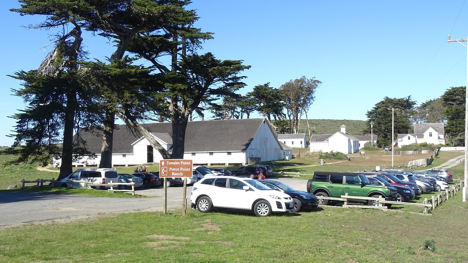 A dirt parking lot with about 2 vehicles with a few trees and white buildings in the background.