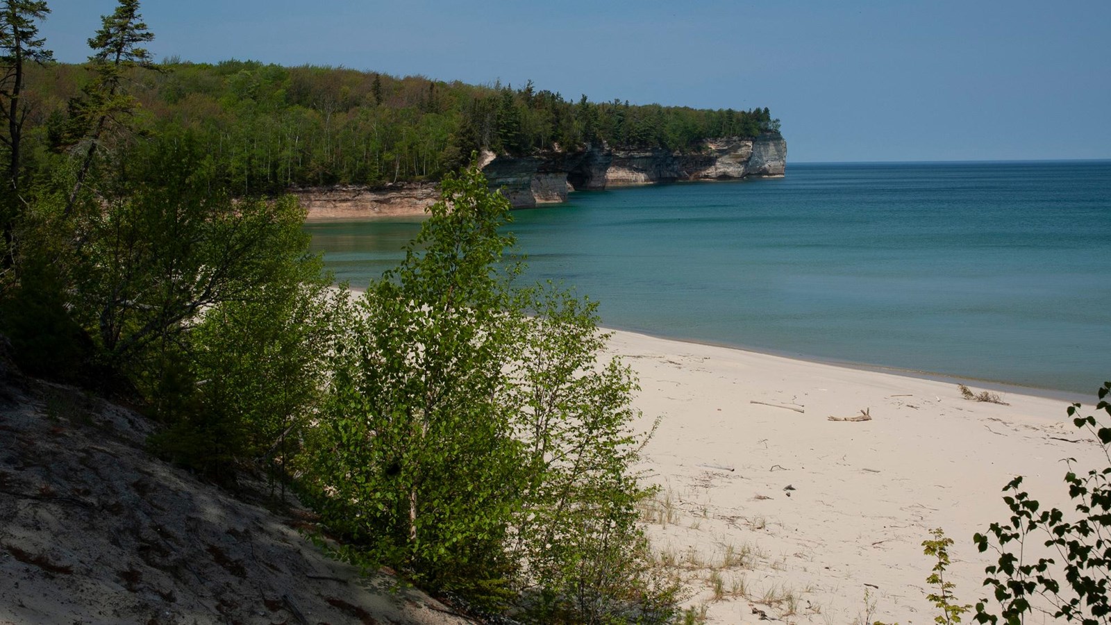 Small beach on Lake Superior. Rock cliffs rise out of the water in the distance. The water is calm.