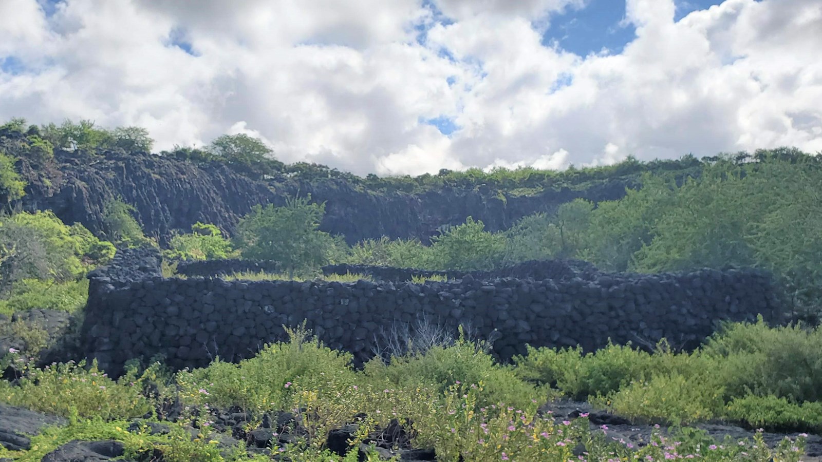 A large enclosure with 6 foot high stacked stone walls lies in front of a lava rock cliffside.