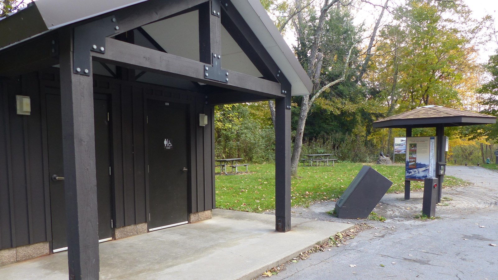 Two door restrooms (left) and a three sided information kiosk along the driveway with a trail (right